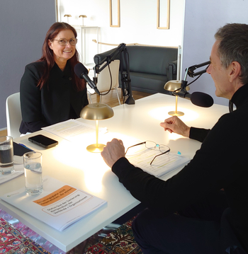 Zwei Personen sitzen sich in einem Podcaststudio gegenüber und führen ein Gespräch vor Mikrofonen.